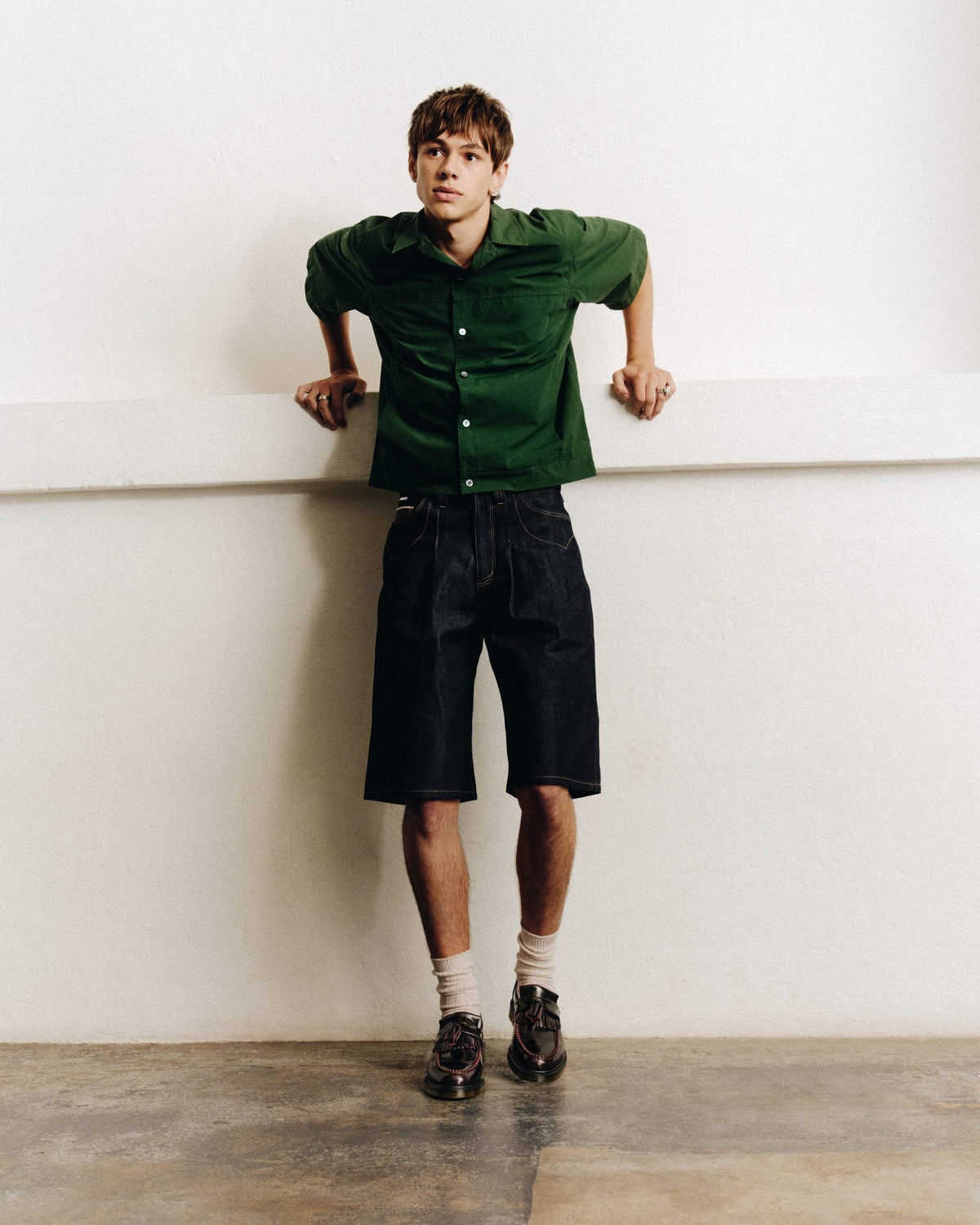 Hombre con camisa verde de manga corta, bermuda de jean larga y mocasines Dr. Martens con borla